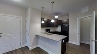 This single-story space showcases a well-appointed kitchen as its centerpiece. The kitchen features rich, dark espresso-toned cabinetry that extends to the upper walls, providing ample storage throughout. A peninsula with a light-toned granite countertop serves as both a prep area and casual dining space, complete with an integrated stainless steel sink. Three pendant lights with classic bell-shaped shades hang above, offering focused task lighting while adding a stylish touch to the space. Black appliances, including a refrigerator, microwave, and range, complement the dark cabinetry for a cohesive, contemporary aesthetic. The kitchen flows seamlessly into the adjacent living area, creating an open-concept feel. Dark hardwood-style flooring runs throughout, providing a warm contrast to the neutral light gray walls and bright white baseboards and trim. The clean lines and neutral color palette lend the space a modern, move-in-ready appeal. Natural light supplements the overhead lighting, contributing to a bright and functional environment overall.