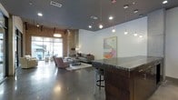 This single-floor space showcases a compelling fusion of industrial and contemporary design elements. The open floor plan centers around polished concrete floors that reflect the ambient lighting, creating a sleek, expansive feel throughout. Exposed brick walls on one side add warm texture and an urban character, contrasting effectively with the smooth white walls adorned with a striking abstract artwork in soft pinks and yellows.

The ceilings are notably high, fitted with a dark finish that enhances the industrial aesthetic, while a combination of recessed lighting and suspended pendant lights provides layered illumination across the space. Large floor-to-ceiling windows along the front facade allow natural light to filter in, connecting the interior to the outside environment.

A substantial stone-topped counter or bar area anchors one side of the space, accompanied by a bar stool, suggesting a functional hospitality or lounge element. The seating area features a neutral-toned sofa, accent chairs in muted tones, and a area rug that defines the lounge zone. The overall ambiance is modern, polished, and welcoming, balancing raw industrial materials with refined furnishings.