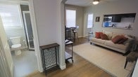 This single-story space showcases a thoughtfully designed open layout that connects multiple living areas through an elegant arched doorway, adding a classic architectural detail to the otherwise contemporary interior. The flooring throughout is rich hardwood, lending warmth and continuity as you move from one area to the next.

The main living area is anchored by a large neutral area rug and a comfortable beige sofa accented with rust-colored throw pillows, creating an inviting and cozy atmosphere. A ceiling fan overhead provides both function and a casual finishing touch. Natural light filters generously into the space through multiple windows fitted with horizontal blinds, keeping the interior bright while allowing for privacy control.

An arched opening leads into a secondary area currently configured as a casual dining or workspace, furnished with bar-height seating and a sleek table. A rolling bar cart near the entryway adds a practical and stylish accent. The neutral gray wall palette throughout ties the spaces together cohesively, while built-in appliances visible along one wall suggest proximity to a kitchen. The overall ambiance is clean, modern, and functional.