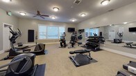 The Greyhawk Townhomes community amenity space is thoughtfully designed to serve residents of all ages, combining a fully outfitted fitness center with a dedicated children's play area. The fitness center is generously sized, featuring neutral gray walls, carpeted flooring, and a textured ceiling with ceiling fans that keep the space comfortable during workouts. Natural light filters in through horizontal-blind-covered windows, complementing the flush-mount lighting throughout. The workout area is equipped with multiple treadmills, elliptical machines, a stationary bike, a stair climber, and a dumbbell rack with a full-length mirror. Wall-mounted televisions provide entertainment, and rubber floor mats protect equipment zones. A posted sign confirms the facility operates 24 hours a day. Adjacent to the fitness area is a vibrant children's playroom decorated with a nautical-themed mural featuring the Greyhawk Townhomes branding. Small chairs, colorful toys, a miniature basketball hoop, and assorted play items fill the room, creating an engaging environment for young residents. Together, these spaces reflect a community-focused design catering to diverse resident needs.