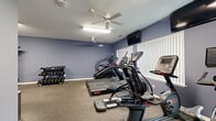 Located at 1688 US-9 in Clifton Park, NY, this fitness center offers a practical and well-equipped exercise space designed to support a variety of workout routines. The room features distinctive blue-painted walls and durable carpet flooring, creating an energetic yet comfortable environment for physical activity.

The cardio section is outfitted with multiple Star Trac treadmills, an elliptical trainer, a stair climber, and a virtual cycling bike, providing a comprehensive range of aerobic options. Several wall-mounted flat-screen TVs are positioned throughout the room, allowing users to stay entertained during workouts. Ceiling fans help maintain air circulation throughout the space.

For strength training, a tiered dumbbell rack stocked with a variety of free weights lines one wall, accompanied by an adjustable workout bench. Additional fitness accessories, including a stability ball and resistance equipment, are available along the perimeter.

A water cooler is conveniently stationed near the interior doors, ensuring hydration is readily accessible. The room includes two doors that connect to adjacent areas of the facility. Natural light filters in through windows dressed with vertical blinds, and ceiling-mounted fluorescent lighting ensures the space remains well-lit at all times. Overall, the fitness center is efficiently organized to maximize usability within its footprint.
