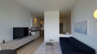 This thoughtfully designed single-floor unit presents a clean, contemporary aesthetic with a highly functional open-plan layout. The living area is bright and airy, featuring light hardwood flooring, smooth white walls, and a crisp white ceiling that together create a sense of openness and calm. A navy blue sofa anchors the living space, complemented by sleek nesting side tables atop a soft area rug, forming a comfortable and stylish seating arrangement. A low-profile media console accommodates a flat-screen television, keeping the space uncluttered and modern. The kitchen is seamlessly integrated into the open floor plan, outfitted with light cabinetry, a stainless steel sink, and stainless steel appliances including a microwave, oven, and dishwasher. Track lighting overhead provides focused illumination throughout the kitchen zone. A partial-height dividing wall elegantly separates the sleeping area from the main living space while preserving the open feel of the unit. The sleeping nook, visible through an open pass-through, features built-in closet storage, adding practical functionality. Overall, the space exudes a minimalist, urban character with a cohesive neutral palette and carefully considered design details.