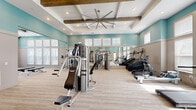 This fitness center offers a generously sized, single-floor workout space designed with both functionality and aesthetics in mind. The architectural design is immediately striking, featuring elevated coffered ceilings accented by rustic wood beams and a large industrial ceiling fan that enhances airflow throughout the open layout. Soft teal walls paired with neutral wainscoting create a bright, energizing atmosphere, while abundant natural light streams in through multiple sets of windows positioned at varying heights along the perimeter walls.

The space is divided into distinct functional zones. A dedicated cardio area houses a row of treadmills, elliptical machines, stair climbers, and a rowing machine, all arranged along a full-length mirrored wall that visually expands the room. Across the floor, a comprehensive strength training section features multi-use cable machines, free weight benches, squat racks, and a tiered dumbbell rack spanning a broad weight range. A separate open zone accommodates functional training, offering TRX suspension equipment, stability balls, medicine balls, and exercise mats. Light-toned flooring—a combination of wood-look panels and low-pile carpet—ties the space together cohesively. Glass-panel doors provide access to an outdoor area, further enhancing the sense of openness.