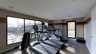 This single-floor fitness center is designed to provide a functional and motivating workout environment. The space is outfitted with a lineup of modern True-brand cardio equipment, including treadmills and elliptical machines, each featuring integrated display screens for an enhanced exercise experience. Large windows span the walls, flooding the room with natural light while offering open views of the fenced outdoor surroundings and neighboring landscape, creating a bright and energizing atmosphere. The neutral-toned walls are accented with a bold horizontal stripe, adding a dynamic visual element to the interior. Recessed lighting in the smooth white ceiling supplements the natural light, ensuring the space is well-lit at all times. The flooring is finished with a combination of carpet and protective equipment mats, providing both comfort and stability during workouts. A dedicated exit door with signage ensures safe and convenient access, while informational boards are mounted near the entrance for resident or member use. The overall design is clean, practical, and welcoming, making it a well-suited facility for daily fitness routines.