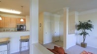 This single-floor residence showcases a thoughtfully designed open-concept layout that seamlessly connects the main living areas. Architectural columns with decorative molded capitals serve as defining structural elements, adding a sense of formality and visual interest to the space while subtly delineating the living and dining zones.

The living area features neutral-toned carpeting and is furnished with warm, earthy-colored seating, contributing to a comfortable and inviting ambiance. A hallway visible from the main living space leads to additional rooms, with multiple white-paneled doors — including a louvered closet door — indicating practical storage and private room access.

The kitchen, visible from the living area, is equipped with dark appliances and features granite or stone countertops. Bar-height seating at the counter provides a casual dining option, and a pendant light fixture overhead adds focused illumination. Light wood cabinetry completes the kitchen's clean, functional aesthetic.

Throughout the space, cream and white walls paired with light-colored trim create a bright, cohesive atmosphere. The combination of carpeted and hard-surface flooring helps define each functional zone within the open floor plan.