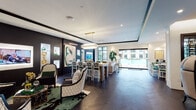 Located at 1916 W Gray St in Houston's desirable River Oaks area, this single-floor amenity space is a masterclass in contemporary interior design. The layout flows seamlessly across several distinct zones, united by rich dark hardwood flooring, tray ceilings with recessed lighting, and a bold black-and-white color palette punctuated by emerald green accents throughout.

The main gathering area features an open-plan arrangement with a stylish lounge seating zone, a central dining table beneath a gleaming brass and crystal chandelier, and curated large-format artwork displayed against dramatic dark-painted accent walls. Floor-to-ceiling black-framed windows and sliding glass doors flood the space with natural light and provide expansive views of the outdoor pool and courtyard beyond.

A private conference or meeting room enclosed in glass walls offers an intimate setting with statement dome-back chairs, a green velvet banquette, and an elegant gold chandelier, all set against textured wall coverings and gallery-style art arrangements.

The kitchen features herringbone-patterned light wood flooring, marble countertops, steel-blue cabinetry with brass hardware, and a large wall-mounted display screen, combining functionality with refined aesthetics.

Additional lounge nooks offer plush neutral sofas, glass-top coffee tables, and framed artwork, all oriented to capture courtyard and pool views. The overall ambiance is sophisticated, curated, and welcoming.