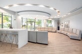 Located at 5191 Dundas St W in Toronto's Etobicoke neighbourhood, this single-floor amenity space is defined by a sophisticated and contemporary design. The centrepiece of the space is a sweeping, curved cream leather sectional sofa arrangement set beneath an elegant circular ceiling feature adorned with brushed gold trim and recessed lighting — a bold architectural statement that draws the eye immediately upon entry. Floor-to-ceiling windows and glass entrance doors flood the space with natural light while offering open views of the surrounding tree-lined streetscape. Light wood-toned flooring runs throughout, reinforcing the warm and welcoming aesthetic. A secondary lounge area features rich brown leather armchairs, curated wall art with an inspirational theme, and tall decorative planters that add a natural, organic element. A dedicated fireplace nook showcases a built-in media and hearth unit with textured wood panelling, open display shelving, and candle accents — creating an intimate retreat within the larger space. A kitchenette with white cabinetry and modern appliances provides functional amenities, while glass-panelled double doors lead to additional corridors and a storage room, ensuring practical utility alongside refined design. The overall ambiance is polished, inviting, and thoughtfully curated.