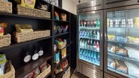 This single-floor retail snack shop is thoughtfully designed to offer a well-organized and convenient shopping experience. The space is anchored by dark-finished open shelving units that line the walls, displaying an extensive selection of packaged snacks, including chips, popcorn, crackers, chocolates, and candy. Woven wicker baskets are used throughout the shelving to neatly categorize and present products, adding a warm, boutique-style aesthetic against the dark shelving frames.

Adjacent to the snack shelving, multiple commercial-grade glass-door refrigerators provide ample cold storage for a broad selection of beverages, including bottled water, energy drinks, sodas, and juices. A separate glass-door freezer unit holds frozen treats and packaged frozen foods, rounding out the product offerings.

The shop is accessed through stylish steel-framed, multi-pane glass doors that lend an upscale, modern feel to the entrance. Light wood flooring in the entry area contrasts with the darker interior tones, creating a welcoming transition into the space. The layout is compact yet efficient, maximizing product visibility and accessibility while maintaining a clean, organized atmosphere throughout.