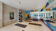 This contemporary fitness studio is designed to energize and inspire. The open floor plan provides generous space for group classes, yoga, and strength training, with light wood flooring that creates a clean, airy foundation throughout. Exposed concrete walls lend an industrial character, beautifully contrasted by vivid, large-scale abstract murals splashed across the walls and ceiling in bold hues of blue, yellow, orange, teal, and red — giving the studio a dynamic, artistic identity.

A full-length mirrored wall expands the visual depth of the room and is framed by the colorful mural accents. The studio is well-equipped with exercise mats, kettlebells, stability balls, balance discs, dumbbell racks, and a Reflect interactive fitness mirror mounted on the concrete wall, offering guided workout sessions.

Natural light floods the space through expansive floor-to-ceiling black-framed glass doors, which open directly to an outdoor courtyard featuring horizontal wood slat fencing and brick surroundings, creating a seamless indoor-outdoor connection. Recessed and track lighting supplements natural illumination throughout the day. A sleek dark storage cabinet with a white countertop provides functional utility near the courtyard entrance, keeping the space organized and ready for use.
