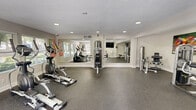 Located at 1408 Knights Trail in Stone Mountain, Georgia, this fitness center offers a well-organized and fully equipped exercise space designed to support a comprehensive workout routine. The single-floor layout provides a generous, open area that accommodates both cardiovascular and strength training equipment without feeling cramped. The cardio section features treadmills and elliptical machines positioned along a row of windows that flood the space with natural light and offer views of the surrounding greenery, creating an energizing atmosphere for endurance training. The strength training area is outfitted with multiple cable and weight machines, allowing users to target a wide range of muscle groups. A large wall mirror enhances the sense of space while allowing users to monitor their form during exercises. The flooring throughout is durable rubber gym matting, providing both safety and comfort underfoot. Recessed lighting complements the natural light, ensuring the space remains bright and well-lit at all times. A wall-mounted flat-screen television is available for entertainment, and a dedicated entry door with an adjacent window provides convenient access and additional ventilation. Vibrant sports-themed artwork adorns the walls, adding an energetic and motivating ambiance to the overall environment.