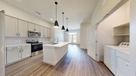 Located at 1431 Firecracker Dr in Buda, TX, this well-designed one-bedroom, one-bathroom unit offers a thoughtfully laid-out single floor of living space. The kitchen is a standout feature, showcasing gray shaker-style cabinetry, a large white quartz center island, stainless steel appliances, and striking black pendant lighting — all set atop wood-look luxury vinyl plank flooring that flows throughout the unit. The bright, open bedroom offers two windows with natural light and outdoor views, along with direct access to two generously sized walk-in closets, each equipped with multi-level shelving and hanging rods for ample storage. The full bathroom impresses with a dual-sink vanity featuring white cabinetry and framed mirrors, open shelving, and a tiled soaking tub and shower combination. A dedicated in-unit laundry closet accommodates a full-size washer and dryer side by side. Clean white walls, modern panel-style doors, and consistent flooring throughout create a cohesive, contemporary aesthetic. The exterior grounds feature lush green landscaping with a wrought iron gate, adding a welcoming curb appeal to this move-in-ready Buda residence.