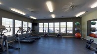 This fitness center offers a dedicated space for exercise and wellness, thoughtfully designed to support a variety of workout routines. The room features dark, durable flooring that provides both stability and easy maintenance, ideal for high-traffic use. Multiple large windows line the walls, allowing ample natural light to fill the space and offering views of the surrounding outdoors, creating an open and energizing atmosphere. Ceiling fans help maintain comfortable airflow throughout the room.

The cardio section is well-appointed, featuring a row of treadmills along one wall, complemented by upright stationary bikes positioned toward the center of the room. Exercise balls are available for stretching and core workouts, adding versatility to the space. A wall-mounted flat-screen television provides entertainment during workouts.

Overhead fluorescent lighting ensures the space remains bright even in low-light conditions. The neutral, dark-toned walls contribute to a focused and functional environment. Overall, this fitness center is a practical and well-organized facility that caters to residents looking to maintain an active lifestyle without leaving the property.