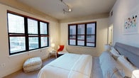 This urban residence offers a thoughtfully designed living space spread across a single floor. The bedroom serves as a central highlight, featuring multiple large windows with warm wood frames and adjustable blinds that frame views of the surrounding city buildings, filling the room with natural light throughout the day. Contemporary track lighting mounted on the ceiling provides flexible, adjustable illumination to complement the natural light. The bedroom accommodates a generously sized bed dressed in crisp white linens, paired with a coordinating upholstered piece that adds a subtle pop of color and texture. A cozy seating nook near the windows offers a quiet spot for reading or relaxing, complete with a small side table and accent chair. The neutral-toned walls and light carpeted flooring create a calm, airy ambiance throughout. The overall aesthetic blends urban industrial character — evident in the bold window surrounds and open city views — with clean, modern furnishings that contribute to a comfortable and functional living environment. The space strikes a balance between city energy and residential tranquility, making it an inviting retreat within an urban setting.