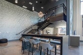 Located at 401 Williams Ave SW in Huntsville, Alabama, this refined two-story commercial space blends modern design with thoughtful detail throughout. The ground floor welcomes visitors with a dramatic double-height lobby, distinguished by soaring ceilings, floor-to-ceiling windows that flood the interior with natural light, and polished concrete floors that add an industrial-chic character. A bold geometric textured accent wall anchors the entry alongside a built-in wooden shelving display, while bistro-style seating arrangements create an inviting atmosphere. A striking steel-railed staircase with warm wood treads and a built-in base cabinet serves as both a functional and architectural focal point, leading to the second floor. Lounge areas on both levels offer flexible social spaces, with plush seating, large-format TVs, and panoramic windows overlooking a well-maintained courtyard and neighboring brick buildings. Intimate nook seating provides quieter retreat spaces, including a cozy corner with floor-to-ceiling glazing and outdoor views. Upstairs, the space transitions to a more refined environment featuring carpeted corridors, an art-forward lounge with velvet furnishings and gold-framed statement artwork, and a polished conference room equipped with a large oval table, pendant lighting, and rich wood cabinetry. Throughout, the design balances contemporary aesthetics with warm, curated touches.