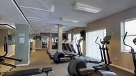 This fitness center offers a comprehensive workout environment designed to accommodate a range of exercise needs. The space is laid out on a single floor, with rows of cardio equipment — including treadmills, elliptical trainers, and stationary bikes — arranged to maximize accessibility and flow. Large windows dressed with vertical blinds line one wall, allowing natural light to filter into the space and creating a bright, open atmosphere.

The ceiling features a distinctive angular, geometric design with recessed and flush-mounted fluorescent lighting panels that ensure consistent illumination throughout. Structural columns are integrated into the layout without significantly disrupting the exercise areas. The neutral beige walls and carpeted flooring contribute to a clean and functional aesthetic.

Toward the back of the space, additional equipment and mirrored walls are visible, extending the sense of depth and providing users with visual feedback during workouts. A wall-mounted television and a clock are also present, adding convenience for exercisers. Overall, the facility presents a practical, well-organized environment suitable for a full cardio and strength training routine.