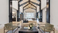 This elegantly designed single-floor common space offers a refined blend of modern and transitional architectural elements. The most striking feature is the dramatic vaulted cathedral ceiling, accentuated by bold, dark exposed structural beams that create a strong visual contrast against the crisp white ceiling and walls. A statement chandelier hangs at the apex, adding a warm, inviting glow to the volume above.

Polished marble tile flooring with subtle diamond inlay details runs throughout, reflecting natural light and contributing to the space's bright, airy atmosphere. Tall, floor-to-ceiling steel-framed windows and glass walls on one side flood the interior with natural light while offering direct views of an outdoor pool area and surrounding stone masonry architecture.

The main lounge area is furnished with a neutral-toned sofa, accent chairs, and a dark coffee table, creating a comfortable yet sophisticated seating arrangement. Adjacent to the lounge, a smaller seating alcove with French doors provides a more intimate setting beneath the same soaring roofline.

A built-in open shelving unit finished in dark tones anchors one wall, adding functional storage and visual depth. A decorative round mirror mounted on a navy accent wall serves as a focal point near a secondary seating and desk area. A kitchenette is also visible, completing this well-appointed, multi-functional communal space.