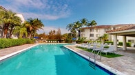 This single-story residence is situated within a vibrant, tropical resort-style community characterized by lush palm tree landscaping and a warm, sun-soaked atmosphere. The standout feature of the property is the expansive outdoor pool area, which serves as the centerpiece of the community's amenities. The generously sized swimming pool is framed by a spacious paved sun deck lined with lounge chairs and shaded by blue umbrellas, creating an inviting environment for relaxation. A covered gazebo provides an additional shaded retreat within the outdoor space, while the surrounding tropical foliage enhances the serene, vacation-like ambiance. The multi-story white stucco buildings with terracotta-tiled rooflines that border the pool area reflect a classic Florida Mediterranean architectural style. The interior of the unit, while not fully captured in available imagery, is contained within a single floor, offering a streamlined and accessible layout. The overall setting conveys a strong sense of resort living, with well-maintained communal outdoor spaces designed for leisure and enjoyment under the Florida sun.