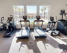 This state-of-the-art fitness center, spanning 630 square feet, offers a comprehensive workout experience. The space is flooded with natural light from large windows, providing views of an outdoor pool area and palm trees. The gym is equipped with a variety of modern exercise machines, including multiple treadmills, ellipticals, and stationary bikes for cardio workouts. A dedicated strength training area features a squat rack, free weights, and weight machines. Mirrored walls enhance the sense of space and allow users to monitor their form. The room's layout is thoughtfully designed, with cardio equipment facing the windows and strength training equipment along the walls. The neutral color scheme, with white walls and carpeted flooring, creates a clean, inviting atmosphere. Recessed lighting and a mounted TV add to the comfortable ambiance. This well-appointed fitness center caters to various exercise preferences, making it an attractive amenity for residents or hotel guests.