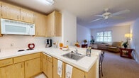 This single-story unit in Clayton, CA, offers a practical and comfortable layout designed with accessibility and ease of living in mind. The kitchen is outfitted with light wood cabinetry providing ample storage, white appliances including a refrigerator, stovetop, and microwave, and white tile flooring. A peninsula counter with a stainless steel sink opens toward the living area, creating a connected, open feel that encourages natural flow between spaces. A ceiling fan overhead serves both the kitchen and adjacent living area.

The living area is bright and welcoming, featuring neutral carpet, white walls, and a window fitted with blinds that allows natural light to filter in. A wall-mounted HVAC unit sits beneath the window, providing climate control. The space is generously sized, offering flexibility for varied furniture arrangements.

The bedroom is carpeted and includes an open closet, a window with natural light, and enough room to accommodate multiple seating pieces comfortably. Vaulted ceiling angles in portions of the space add visual interest and an airy quality.

The bathroom is designed with accessibility as a priority, featuring multiple stainless steel grab bars, a wide layout with tile flooring, and a light wood vanity with storage. The overall unit maintains a clean, bright, and functional character throughout.