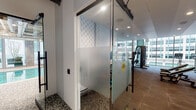 Located in the heart of Chicago at 159 N Wabash Ave, this amenity floor delivers a comprehensive suite of lifestyle and wellness facilities within a modern, industrial-inspired setting. Exposed concrete ceilings, visible ductwork, and floor-to-ceiling glass walls define the architectural character throughout the space, blending raw industrial elements with polished contemporary finishes.

The fitness center is a standout feature, divided into distinct zones. A cardio area lines the expansive glass perimeter, offering cycling bikes, a rowing machine, and a cable rack against sweeping views of the Chicago skyline. An adjacent strength training zone houses multiple cable and weight machines alongside medicine ball storage. A separate exercise studio with large-screen displays and frosted glass partitions provides a dedicated space for guided or independent training sessions.

The wet area features mosaic tile floors and walls, with access-controlled entry via a key fob panel, complementing the facility's modern security design.

The lounge area offers a warm, social atmosphere with wood-look flooring, curated artwork, wall-mounted TVs, a wet bar with beverage refrigeration, and a ping pong table. Sliding glass doors open directly onto a furnished outdoor terrace complete with a pergola, grill station, and checkerboard tile patio — offering a rare urban outdoor retreat amid Chicago's vibrant Loop neighborhood.