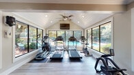 Located at 705 Powers Ferry Rd SE in Marietta, Georgia, this single-floor fitness center is thoughtfully designed to provide a bright, welcoming environment for exercise. The main workout area is anchored by a striking vaulted, shiplap ceiling with recessed lighting and a ceiling fan, creating an airy, open atmosphere. Floor-to-ceiling and oversized windows line the walls, flooding the space with natural light and offering direct views of the outdoor swimming pool and surrounding mature trees — an inspiring backdrop for cardio sessions. The cardio zone is equipped with multiple treadmills, elliptical trainers, upright and recumbent bikes, and a rowing machine, all positioned to take advantage of the poolside views. A dedicated free-weight and strength corridor runs along one side of the facility, featuring large windows that overlook the landscaped grounds and adjacent sport courts. Wall-mounted mirrors and a punching bag round out the strength training offerings. The space also includes a central area housing cable and functional training machines. Light wood-look flooring flows throughout, complementing the neutral warm-gray walls and crisp white trim. Separate men's and women's restrooms are conveniently located within the facility, accented by bold black doors that add a modern contrast to the clean, classic interior design.
