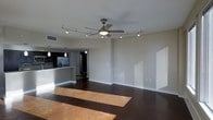 This single-floor space offers a thoughtfully designed open-concept layout that seamlessly connects the living area to the kitchen. The living space is anchored by rich, dark hardwood flooring that contrasts beautifully with the clean, neutral-toned walls, creating a contemporary and inviting atmosphere. Natural light streams in, casting warm patches across the floor and highlighting the spaciousness of the room.

Overhead, a stylish ceiling fan with an integrated light fixture sits at the center of the living area, complemented by a curved track lighting system that provides flexible, directed illumination throughout the space. The combination of these lighting elements ensures the room is both functional and well-lit at any time of day.

The kitchen features a modern design with dark cabinetry, a stainless steel refrigerator, and a raised breakfast bar that provides a natural division between the cooking and living areas. Pendant lights hang above the kitchen counter, adding a refined touch to the culinary space. The layout allows for easy flow between areas, making the space ideal for both everyday living and entertaining. Overall, the space presents a clean, minimalist aesthetic with quality finishes throughout.