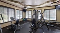 This fitness center at 29675 N North Valley Pkwy in Phoenix, Arizona offers a well-equipped workout facility designed to support a comprehensive exercise routine. The space features a distinctive tray ceiling with elegant crown molding details, overhead ceiling fans for enhanced air circulation, and recessed lighting that keeps the environment bright and welcoming. Dark wood-trimmed windows fitted with horizontal blinds line the walls, allowing natural light to filter in while maintaining privacy and providing views of the surrounding exterior. Entry is through a set of classic French doors with dark wood framing, adding an architectural elegance to the space. The flooring consists of durable dark rubber matting suited for high-traffic fitness use. Cardio equipment includes multiple treadmills and elliptical machines, while the strength-training area is outfitted with a multi-station cable machine, a Smith machine, a weight bench, a full dumbbell rack, and stability balls of various sizes. Full-length mirrors are strategically placed to assist with form and to visually expand the room. A wall-mounted television provides entertainment during workouts. The overall ambiance is clean, functional, and thoughtfully designed, striking a balance between practicality and refined aesthetic detail.
