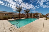 Located at 5821 W McDowell Rd in Phoenix, Arizona, this single-level outdoor amenity space offers a resort-style pool experience designed for relaxation and leisure. The centerpiece is a generously sized rectangular swimming pool with clear turquoise water, accessible via stainless steel entry rails and marked with depth indicators for safety. The pool deck is finished in a light-toned concrete surface that surrounds the water on all sides, providing ample space for poolside activity. Tall, mature palm trees are strategically placed throughout the area, offering natural shade and a distinctly Southwestern aesthetic. The lounge area features navy blue chaise lounges arranged on a well-maintained artificial turf lawn, complemented by striped navy and white patio umbrellas and small white accent tables. A covered pergola-style structure with a Spanish tile roof provides a shaded gathering space furnished with outdoor seating, ideal for dining or socializing. The surrounding buildings display classic Spanish Colonial Revival architecture, characterized by stucco exteriors, arched entryways, tile roofing, and stone accent detailing. Desert-appropriate landscaping with rock beds and native shrubs frames the perimeter. The space is enclosed by an iron fence, ensuring privacy while preserving open sightlines beneath Arizona's expansive sky.