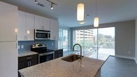 Located at 202 N West St in the heart of Raleigh, NC, this single-floor unit offers a thoughtfully designed open-concept layout filled with contemporary finishes and abundant natural light. The kitchen serves as a focal point of the space, anchored by a large granite-topped island with an undermount sink and a sleek gooseneck faucet. White cabinetry with clean-lined hardware contrasts beautifully with dark lower cabinets and a striking blue mosaic tile backsplash. Stainless steel appliances, including a range and over-the-range microwave, complete the modern culinary setup. Track lighting overhead provides ample task illumination throughout the kitchen. Perhaps the most impressive feature of the space is the expansive floor-to-ceiling windows that line the living area, flooding the interior with natural light and offering open views of the surrounding Raleigh cityscape and lush tree canopy. These large glazed panels create a seamless visual connection between the interior and the outdoors, lending the space an airy, bright ambiance. The overall aesthetic is sleek and urban, with a neutral color palette that allows the architectural details and city views to take center stage, making this a compelling space in one of Raleigh's vibrant downtown neighborhoods.