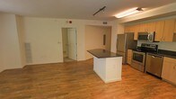 This single-floor space is designed with an open-concept layout that seamlessly connects the main living area to the kitchen. Warm-toned hardwood flooring extends throughout, lending continuity and a welcoming feel to the interior. The kitchen is well-equipped with stainless steel appliances, including a full-sized refrigerator and range, complemented by light wood cabinetry that provides ample storage. A central island with a dark countertop serves as a natural dividing point between the kitchen and the open living space, offering additional workspace or casual seating. Track lighting and flush-mounted fixtures illuminate the space effectively, while a textured ceiling adds subtle visual interest overhead. The neutral, light-colored walls keep the interior feeling bright and versatile. A doorway visible from the main area leads to an adjoining room, suggesting a functional multi-room layout. The open floor plan allows for flexible furniture arrangement, making the space adaptable to a range of uses. Overall, the interior maintains a clean, practical aesthetic suited to everyday living.
