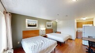 This compact single-floor unit in Baton Rouge, Louisiana, is designed for efficient, functional living. The spacious main room accommodates two full-sized beds with warm wood-toned headboards, flanked by a shared nightstand and wall-mounted lamp. Sage green accent walls are complemented by neutral tones and framed landscape artwork, creating a calm, understated atmosphere. Hardwood-style flooring runs throughout, adding warmth and continuity across the space. A window with curtains allows natural light to filter into the room. The kitchenette is efficiently arranged with warm wood cabinetry, a speckled stone-look countertop, an electric coil cooktop, an over-the-range microwave, and a sink — providing essential cooking and food preparation capabilities. The bathroom is straightforward and functional, featuring a shower-tub combination with a white curtain, a toilet, hardwood-style flooring consistent with the rest of the unit, and a vanity with a stone-look countertop. The textured ceiling and efficient layout reflect a practical design approach, making this unit well-suited for both short-term and extended stays requiring essential day-to-day amenities.