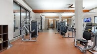 Located at 3000 Forrest Walk in Roswell, Georgia, this well-appointed fitness center offers a generously sized, single-floor exercise facility designed to support a comprehensive workout experience. The space is defined by its striking architectural details, including bold orange ceiling beams that contrast with crisp white walls, exposed brick accents, and warm wood paneling that lend the room a modern yet inviting character.

The main exercise area is thoughtfully divided into distinct zones. A dedicated strength training section hosts a wide array of cable machines and selectorized equipment arranged in an open layout, allowing for easy movement between stations. A free weights area features a full dumbbell rack accompanied by mirrors that visually expand the space and support proper form. The cardio zone includes elliptical trainers and other aerobic equipment, complemented by a wall-mounted television for entertainment during workouts.

Durable rubber flooring — terracotta-toned in the main workout area and a neutral speckled finish near the entrance — provides traction and support throughout. Ceiling fans and overhead lighting ensure the space remains comfortable and well-lit. A built-in dual water fountain station near the entry offers convenient hydration access. Storage cubbies near the entrance provide practical organization. Large windows allow natural light to filter in, enhancing the overall energizing atmosphere of this fully equipped fitness facility.