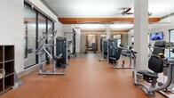 Located at 3000 Forrest Walk in Roswell, Georgia, this well-appointed fitness center offers a generously sized, single-floor exercise facility designed to support a comprehensive workout experience. The space is defined by its striking architectural details, including bold orange ceiling beams that contrast with crisp white walls, exposed brick accents, and warm wood paneling that lend the room a modern yet inviting character.

The main exercise area is thoughtfully divided into distinct zones. A dedicated strength training section hosts a wide array of cable machines and selectorized equipment arranged in an open layout, allowing for easy movement between stations. A free weights area features a full dumbbell rack accompanied by mirrors that visually expand the space and support proper form. The cardio zone includes elliptical trainers and other aerobic equipment, complemented by a wall-mounted television for entertainment during workouts.

Durable rubber flooring — terracotta-toned in the main workout area and a neutral speckled finish near the entrance — provides traction and support throughout. Ceiling fans and overhead lighting ensure the space remains comfortable and well-lit. A built-in dual water fountain station near the entry offers convenient hydration access. Storage cubbies near the entrance provide practical organization. Large windows allow natural light to filter in, enhancing the overall energizing atmosphere of this fully equipped fitness facility.