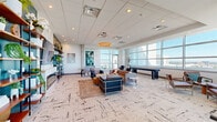 Located at 101 Lake Ave in the heart of Orlando, FL, this sophisticated high-rise amenity lounge offers an impressive blend of modern design and urban living. Floor-to-ceiling windows wrap around the space, flooding the interior with abundant natural light and framing sweeping panoramic views of the Orlando skyline and surrounding cityscape. The open, single-floor layout is thoughtfully organized into distinct zones, each serving a unique purpose while maintaining a cohesive, contemporary aesthetic. A recreational area features a foosball table and a shuffleboard table, perfect for social gatherings, while a comfortable lounge zone is anchored by stylish modular seating, a circular pendant light, and curated artwork that adds a vibrant pop of color. A dedicated nook with a glass-top table and a bold teal shelving display wall provides an intimate setting for smaller meetings or casual conversations. The textured, patterned carpeting ties the space together throughout. Concrete columns punctuate the interior, lending an industrial character that complements the clean white ceilings and recessed lighting. A private balcony extends the experience outdoors, offering unobstructed, breathtaking views of downtown Orlando under open skies — a true highlight of this elevated urban retreat.