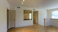 This single-story space showcases a clean, open layout designed for easy living and functional flow. The main living area features light-toned hardwood flooring that extends throughout, contributing to a warm and cohesive aesthetic. White walls and a neutral color palette create a bright, airy atmosphere, enhanced by overhead track lighting that provides focused illumination.

A defining architectural feature is the pass-through kitchen window, which opens from the kitchen into the main living area. This cut-out is framed by a dark countertop, creating a visual connection between the two spaces while maintaining a sense of defined zones. The kitchen is partially visible through the opening, revealing cabinetry, additional track lighting, and ample counter space.

An open doorway on the right side of the main living area leads to additional rooms, offering a glimpse of continued hardwood flooring and neutral finishes beyond. A decorative column element is visible near the right wall, adding a subtle architectural detail. HVAC vents are positioned thoughtfully throughout the ceiling, ensuring comfortable climate control. The overall ambiance is clean, minimal, and move-in ready.