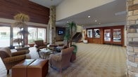 This clubhouse offers a thoughtfully designed single-floor layout that balances elegance with functionality. The expansive main lounge serves as the heart of the space, featuring soaring ceilings with rich wood paneling, a striking stone fireplace column, and patterned carpeting throughout. Comfortable seating arrangements centered around glass coffee tables create inviting areas for socializing, while large windows and glass doors flood the interior with natural light and views of the surrounding landscape. A dedicated sunroom off the main lounge is lined with wide windows and lattice transoms, furnished with plush armchairs, a sectional sofa, and a bistro-style dining table — ideal for casual relaxation or intimate gatherings. The reception area includes a curved granite-topped desk with warm wood cabinetry and pass-through windows that connect to an adjacent office, ensuring a welcoming and organized point of entry. The fully equipped kitchen features light wood cabinetry, tile countertops, black appliances, and tile flooring, accommodating events and catering needs with ease. A separate dining nook within the kitchen area provides additional seating. Throughout the clubhouse, warm earth tones, natural wood accents, and carefully selected furnishings contribute to a cohesive and inviting atmosphere.