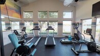 Located at 1220 Crawford Ct in Granbury, Texas, this single-floor fitness center is thoughtfully designed to offer a comprehensive workout environment with an inspiring backdrop. The space features vaulted ceilings that create an open, airy atmosphere, enhanced by large banks of windows that flood the room with natural light and frame picturesque views of the surrounding greenery and adjacent waterway.

The cardio zone is well-stocked with multiple treadmills, elliptical trainers, a stationary bike, and a rowing machine, all positioned to take advantage of the scenic water views. A wall-mounted flat-screen television is also present for entertainment during workouts. The strength training area includes a cable machine, a weight bench, and additional resistance equipment, complemented by a full-length mirror wall and a motivational sunrise mural featuring a runner — reinforcing the fitness-focused atmosphere.

The flooring throughout is durable rubber matting suited for high-activity use. Teal and gold horizontal accent stripes add a consistent decorative touch along the walls. A glass-paneled door connects the fitness room to an adjacent interior space, while a separate entrance opens directly to a covered stone-columned patio overlooking the water, seamlessly blending indoor exercise with the natural beauty of the Granbury lakeside setting.