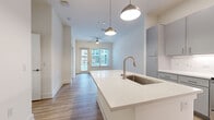 Located at 1251 Ray Charles Blvd in Tampa, Florida, this well-designed one-bedroom, one-bathroom residence offers a thoughtfully utilized layout across a single floor. The open-concept kitchen and dining area serves as the heart of the home, featuring sleek grey cabinetry, stainless steel appliances including a range, microwave, and refrigerator, and bright white quartz countertops. A central kitchen island with an undermount sink and stylish pendant lighting adds both functionality and visual appeal. Light hardwood-style flooring flows throughout the kitchen, dining, and living areas, contributing to a clean, modern aesthetic. The living area is bathed in natural light through large windows and a glass door, complemented by a ceiling fan overhead. The bedroom is a comfortable retreat with soft carpeting underfoot, a bright window with exterior views, and access to a dedicated closet. The full bathroom is finished with a dual-sink vanity featuring grey cabinetry and brushed nickel hardware, a large mirror, and a full-size soaking tub with tile surround. Light wood-tone flooring ties the bathroom's design together. Neutral tones and contemporary finishes throughout create a bright, cohesive living environment.
