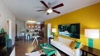 This well-appointed 2-bedroom, 2-bathroom single-floor residence offers a thoughtfully designed open-concept layout that seamlessly connects the living, dining, and kitchen areas. The living room makes a striking first impression with a bold yellow accent wall serving as a dramatic backdrop for a large-scale landscape painting. Rich hardwood-style flooring runs throughout, adding warmth and continuity to the space. A stylish ceiling fan anchors the living area, which is furnished with a clean-lined white sofa accented by teal and gold throw pillows, and complemented by a distinctive teal coffee table atop a cowhide rug. The dining area features a compact round table with modern seating, ideal for casual meals. The kitchen is open to the main living space and showcases dark wood cabinetry, stainless steel appliances, and a peninsula with pendant lighting and bar seating — perfect for entertaining. An arched architectural detail adds a classic design touch to the transition between rooms. The overall ambiance is energetic yet inviting, blending contemporary furnishings with vibrant color choices to create a lively and functional living environment.
