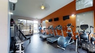 This single-floor fitness center is designed to support a well-rounded workout experience. The space is immediately defined by a striking burnt-orange accent wall that adds energy and vibrancy to the room. Mounted along this wall are multiple flat-screen TVs, allowing users to stay entertained during cardio sessions. The cardio area is equipped with a row of treadmills and an elliptical machine, all positioned to face the screens for a comfortable workout experience. Natural light filters into the space through a large sliding glass door and adjacent windows, creating a bright and welcoming atmosphere. Near the entrance, a dedicated free weights and strength training zone includes a weight bench, a rack of medicine balls, and a stability ball, offering versatility for a range of exercises. The flooring throughout is a durable, dark rubberized surface, practical for high-impact activities and easy maintenance. The layout is thoughtfully organized to maximize usable floor space while keeping equipment accessible. The overall ambiance is functional, motivating, and well-maintained, making it a capable facility for daily fitness routines.