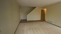 This two-floor space offers a straightforward and functional layout. The main living area is finished with light-toned carpeting that covers a generous portion of the floor, transitioning to a hardwood-style surface near the base of the staircase and adjacent areas. The staircase, positioned toward the back of the room, features clean angular lines and rises to the upper floor, with the underside forming a distinctive triangular profile along the wall. Neutral, light-toned walls throughout the space contribute to a bright and understated ambiance. A doorway near the staircase leads to an additional room or hallway, hinting at further living or utility space beyond the main area. A second door is visible along the left wall, providing additional access points within the layout. Recessed ventilation fixtures are visible in the ceiling, suggesting a central climate system. The overall design is simple and practical, with minimal ornamentation, making it a versatile space adaptable to a variety of furnishing and décor styles. The two-floor configuration provides a clear separation between living areas across levels.