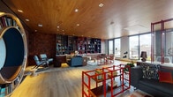 This sophisticated single-floor space showcases a carefully curated interior defined by a seamless blend of warm materials and bold design elements. The ceiling is clad entirely in rich wood paneling, complemented by recessed lighting that casts an inviting, even glow across the open layout.

A standout circular bookcase mounted on one wall serves as a dramatic focal point, while floor-to-ceiling built-in shelving in dark walnut lines another wall, displaying books, decorative objects, and curated collectibles. Bold red powder-coated steel shelving units are distributed throughout the space, adding striking industrial contrast to the warmer tones.

The living area features a large sectional sofa, accent chairs in muted blue-gray tones, and layered area rugs that define distinct seating zones. A textured, patchwork-style feature wall in earthy copper and brown tones anchors the lounge area with visual depth.

Floor-to-ceiling windows along one side flood the interior with natural light while offering broad views of the surrounding cityscape. Light-toned hardwood flooring runs throughout, unifying the space. The overall ambiance is eclectic yet refined, balancing industrial accents with cozy, lived-in warmth.