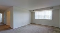 This single-floor space offers a clean, functional layout designed with simplicity and comfort in mind. The main living area is generously proportioned, featuring wall-to-wall carpeting in a neutral tone that adds warmth underfoot while complementing the crisp white walls throughout. A large picture window fitted with vertical blinds serves as a focal point, allowing abundant natural light to flood the interior and creating a bright, airy atmosphere. The window also provides a view of the surrounding exterior, enhancing the sense of openness within the room. The ceiling is smooth and finished with a consistent white tone, contributing to the overall clean aesthetic of the space. Subtle architectural details, including a slightly recessed ceiling beam, add mild visual interest without disrupting the minimalist character of the room. Ventilation is provided via ceiling-mounted vents, ensuring year-round climate comfort. The neutral color palette and uncluttered layout make this space highly adaptable, offering a versatile canvas well-suited for a variety of furnishing arrangements and interior styles. Overall, the space presents a straightforward, light-filled environment that emphasizes practicality and ease of use.