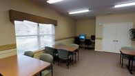 This single-floor space functions as a combined meeting and computer room, offering a practical layout suited for both collaborative and independent use. The room is finished with warm beige walls accented by a decorative wallpaper border that runs along the mid-wall, adding a subtle touch of character to the otherwise functional interior. Carpeted flooring in a neutral tone contributes to a quiet, comfortable atmosphere throughout.

A central rectangular table surrounded by padded, cushioned chairs in muted green serves as the primary meeting area, accommodating small group gatherings or study sessions. Natural light enters through a large window dressed with horizontal blinds and a fabric valance, brightening the space during daytime hours.

Along the far wall, a built-in desk area hosts multiple computer workstations equipped with monitors and office chairs, providing dedicated space for individual computer use. Fluorescent overhead lighting ensures consistent illumination throughout the room. A white door on the far wall offers access to an adjoining area. Overall, the space is clean, organized, and well-suited for community or office use.