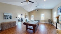 This single-story amenity center is thoughtfully designed to offer a variety of recreational and wellness spaces. The game room serves as a central highlight, featuring a full-size pool table set atop warm hardwood-style flooring beneath soaring vaulted ceilings fitted with ceiling fans and recessed lighting. Large windows allow ample natural light to fill the room, creating a bright and welcoming atmosphere. A wall-mounted cue rack and bar-height seating add functional touches to the space.

Adjacent to the game room is a comfortable lounge area with neutral-toned sofas, decorative wall art, and vaulted ceilings that enhance the sense of openness. This area provides a relaxed setting for socializing or unwinding, with direct access to the fitness center.

The fitness center is generously sized and well-lit, with multiple windows that bring in natural light from multiple sides. A bold terra cotta accent wall adds visual character to the room. Equipment includes treadmills, ellipticals, stationary bikes, a cable weight machine, and a free weight rack with dumbbells. Durable rubber flooring, ceiling fans, and wall-mounted televisions make this an inviting and functional workout environment. French doors connect the fitness center to the lounge, ensuring a seamless flow throughout the facility.