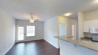 This single-floor space showcases an open-concept layout that seamlessly connects the kitchen and living areas. The kitchen features a peninsula with a granite countertop, providing both functional workspace and a natural divider between the two zones. The living area is brightened by a large window that allows ample natural light to fill the room, complemented by a ceiling fan with a built-in light fixture overhead. Dark hardwood-style flooring runs throughout, creating a rich contrast against the neutral, light gray walls and white trim. The textured ceiling adds a subtle architectural detail to the space. A flush-mount ceiling light provides additional illumination near the entryway or hallway area. Doorways visible in the background suggest access to additional rooms, maintaining a practical and functional flow throughout the unit. The overall ambiance is clean, modern, and move-in ready, with a neutral palette that offers flexibility for a variety of interior design styles. White baseboards and clean lines throughout contribute to the space's polished, well-maintained appearance.