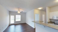 This single-floor space showcases an open-concept layout that seamlessly connects the kitchen and living areas. The kitchen features a peninsula with a granite countertop, providing both functional workspace and a natural divider between the two zones. The living area is brightened by a large window that allows ample natural light to fill the room, complemented by a ceiling fan with a built-in light fixture overhead. Dark hardwood-style flooring runs throughout, creating a rich contrast against the neutral, light gray walls and white trim. The textured ceiling adds a subtle architectural detail to the space. A flush-mount ceiling light provides additional illumination near the entryway or hallway area. Doorways visible in the background suggest access to additional rooms, maintaining a practical and functional flow throughout the unit. The overall ambiance is clean, modern, and move-in ready, with a neutral palette that offers flexibility for a variety of interior design styles. White baseboards and clean lines throughout contribute to the space's polished, well-maintained appearance.