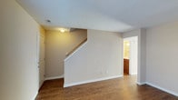 This two-floor residence offers a straightforward and functional layout designed for comfortable everyday living. The main level showcases durable hardwood-style flooring in a rich, warm tone that runs throughout the open living area, creating a cohesive and inviting atmosphere. Neutral beige walls are complemented by crisp white baseboards and trim, lending the space a clean, well-maintained appearance.

A prominent staircase rises from the main living area, featuring a classic wooden handrail and providing direct access to the upper floor. The staircase is positioned centrally, serving as a natural visual anchor within the space. A textured ceiling adds a subtle design detail overhead, while a ceiling-mounted light fixture provides ambient illumination.

Adjacent to the main living area, a doorway leads to a bathroom that is visible in the background, featuring warm wood cabinetry and well-lit vanity lighting. The entry door, finished in white, opens directly into the main living space, offering convenient and immediate access to the home's interior.

The upper floor, accessible via the staircase, expands the overall living capacity of the home. The layout is practical and efficient, with each area flowing naturally into the next, making this a well-organized two-level space suited for a variety of living arrangements.