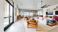 Located in Phoenix, AZ, this thoughtfully designed single-floor community space offers an open and inviting layout that seamlessly connects a living room, dining area, and kitchen. The living room anchors the space with a large, statement ceiling fan, dual wall-mounted televisions above a floating wood credenza, and a display shelf for décor. Comfortable seating including a plush white sofa, colorful accent chairs, and warm wood armchairs create an inviting lounge atmosphere, all centered around a round pedestal coffee table atop a woven area rug. The dining area is bright and airy, featuring multiple round tables with modern chairs, bold geometric-patterned wallcovering, and a striking multi-globe pendant light fixture. A charming built-in banquette with vibrant striped upholstery and a round café table offers an intimate seating nook. Large sliding glass doors flood the dining area with natural light and provide direct access to an outdoor patio. The kitchen is sleek and functional, outfitted with white shaker cabinetry, white quartz countertops, a stainless steel French door refrigerator, and a bold terracotta geometric tile backsplash that adds personality and warmth. Light tile flooring flows throughout the entire space, tying each zone together cohesively.