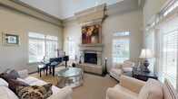 Located at 3015–3069 E 51st St in Tulsa, Oklahoma, this single-story community space is defined by its impressive architectural design and refined interior details. The centerpiece of the main gathering area is a dramatic floor-to-ceiling stone fireplace with an ornate mantel, flanked by large windows fitted with plantation shutters that allow generous natural light to fill the room. Soaring ceilings with crown molding and a ceiling fan enhance the sense of openness and grandeur throughout the space.

The main lounge area is comfortably furnished with upholstered seating arranged around a glass-top coffee table, creating an inviting setting for socializing or relaxation. A grand piano positioned near the windows adds a distinguished, elegant touch. An adjacent dining area features a large rectangular table with cushioned chairs and direct access to an outdoor patio through a glass-paneled door, offering views of the surrounding greenery. Transom windows throughout connect the dining and lounge areas visually, maintaining a cohesive, open flow.

The flooring features a neutral patterned carpet that complements the warm, taupe-toned walls. Overall, the space presents a polished, welcoming ambiance suited for gatherings, events, or everyday community use.